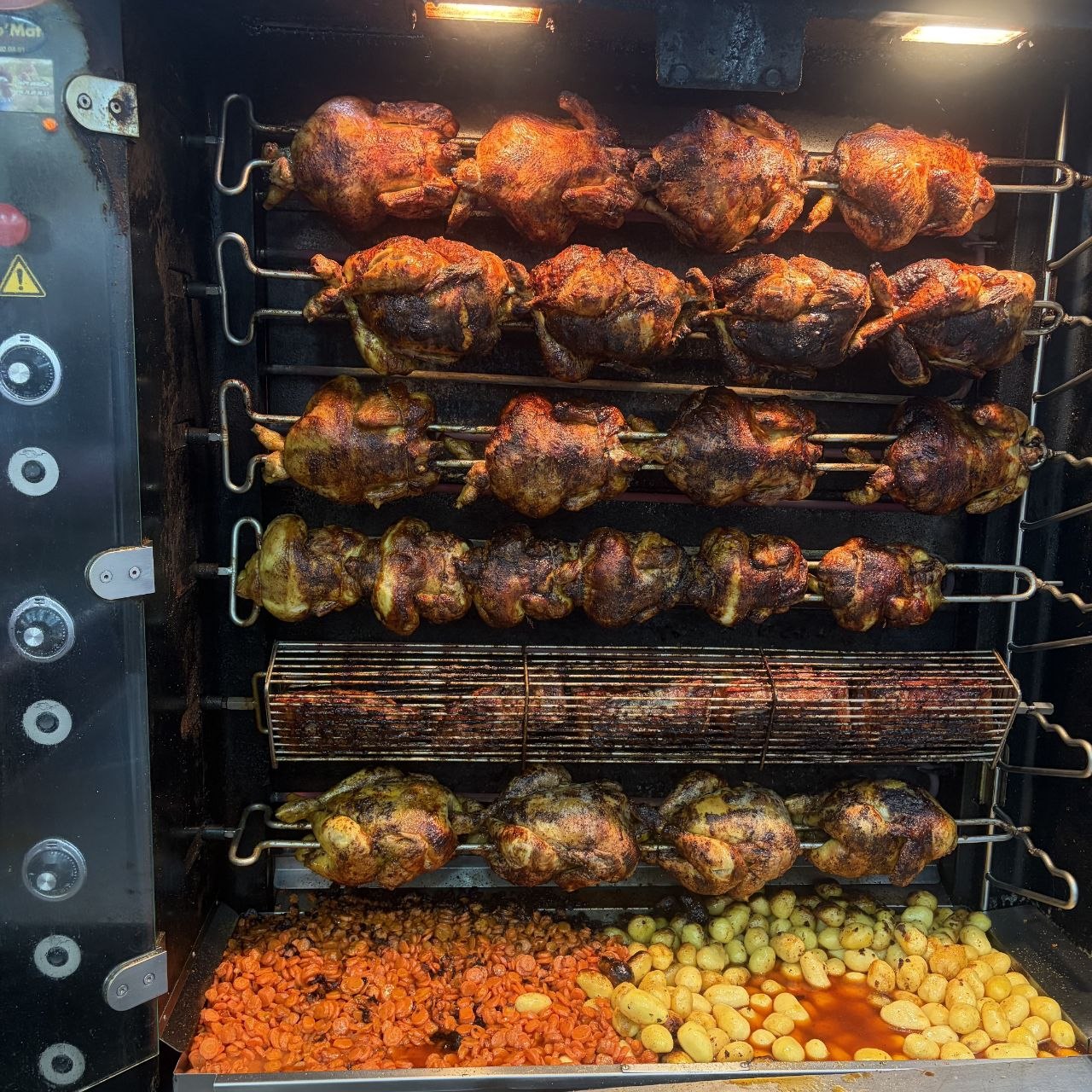 La rôtissoire de Chez Dave : six broches de poulets dorés au-dessus du bac de pommes de terre et de carottes.